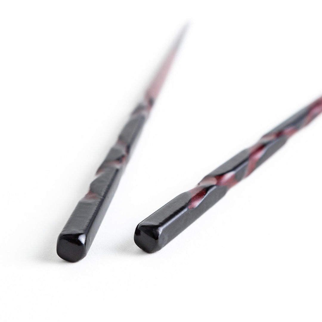 A pair of twisted wooden chopsticks in a dark red and black color gradient.