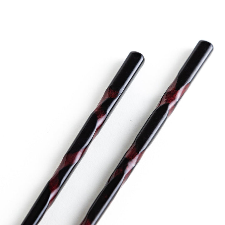 A pair of twisted wooden chopsticks in a dark red and black color gradient.