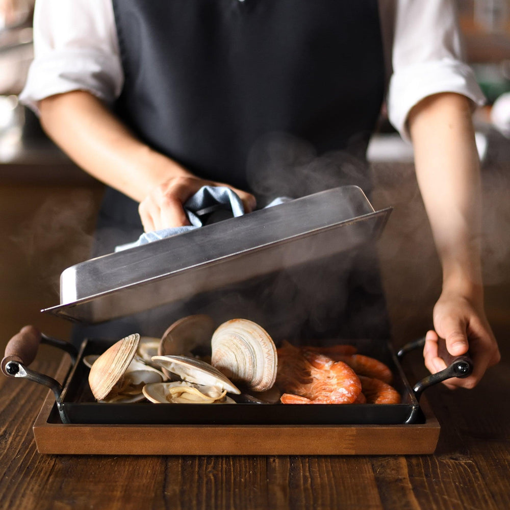 Person lifting a cast iron griddle lid, revealing steaming seafood.