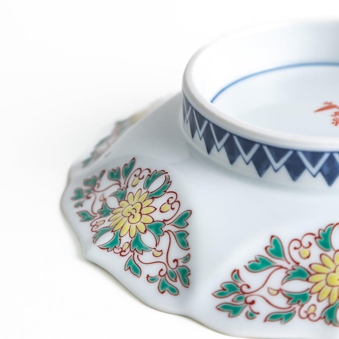 A blue dessert plate with a geometric pattern in the center, accented by ornate floral designs in red, green, and gold on the scalloped rim.