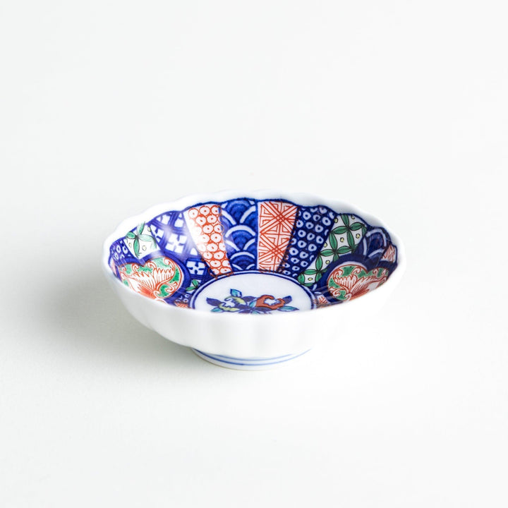 A small condiment bowl with a white interior and a scalloped edge, decorated with a central floral motif and colorful geometric patterns in blue, red, and green.