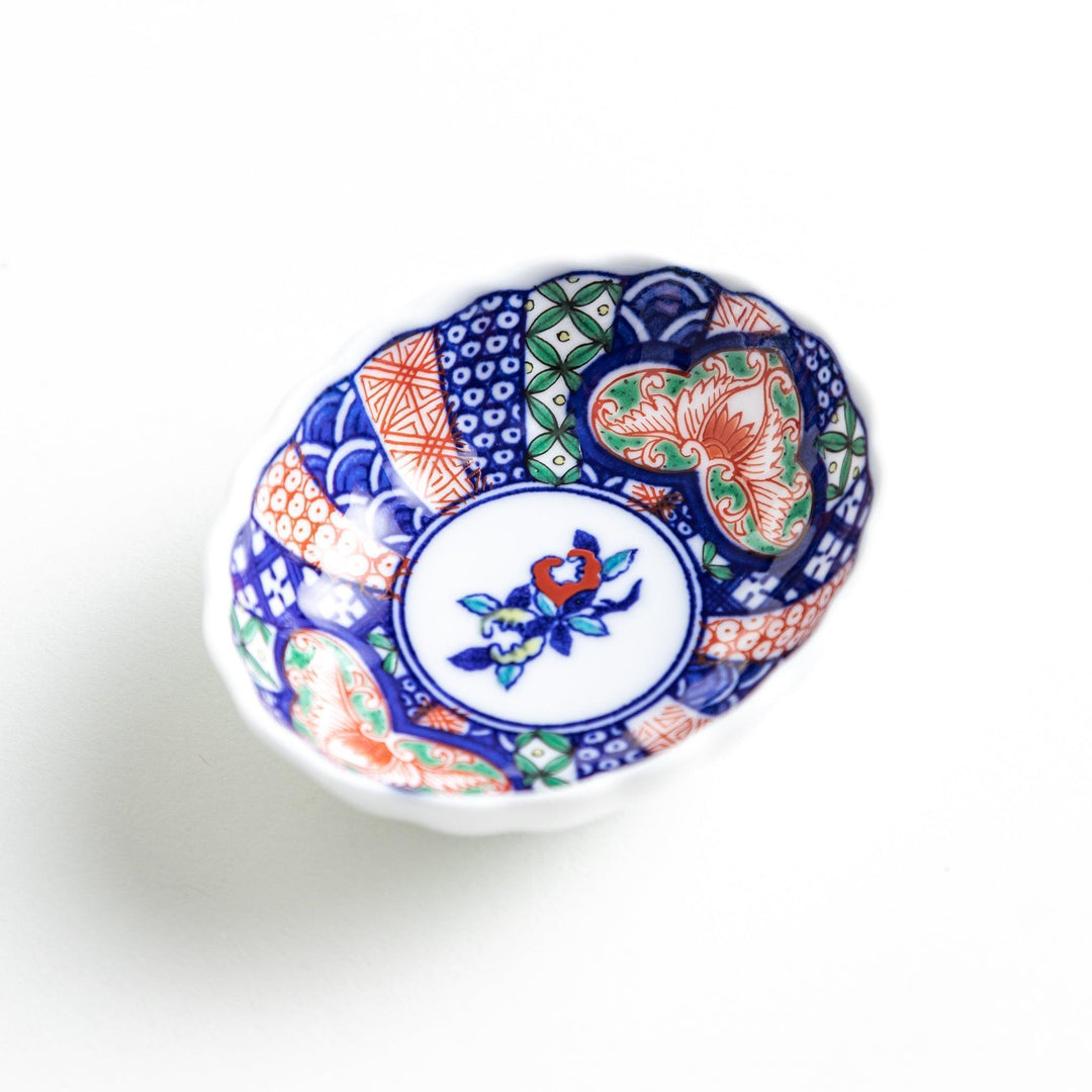 A small condiment bowl with a white interior and a scalloped edge, decorated with a central floral motif and colorful geometric patterns in blue, red, and green.