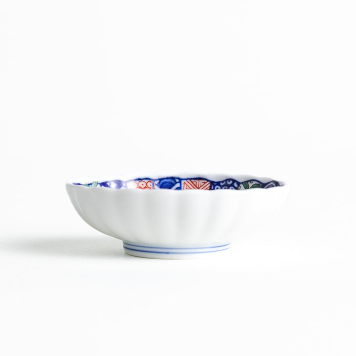 A small condiment bowl with a white interior and a scalloped edge, decorated with a central floral motif and colorful geometric patterns in blue, red, and green.