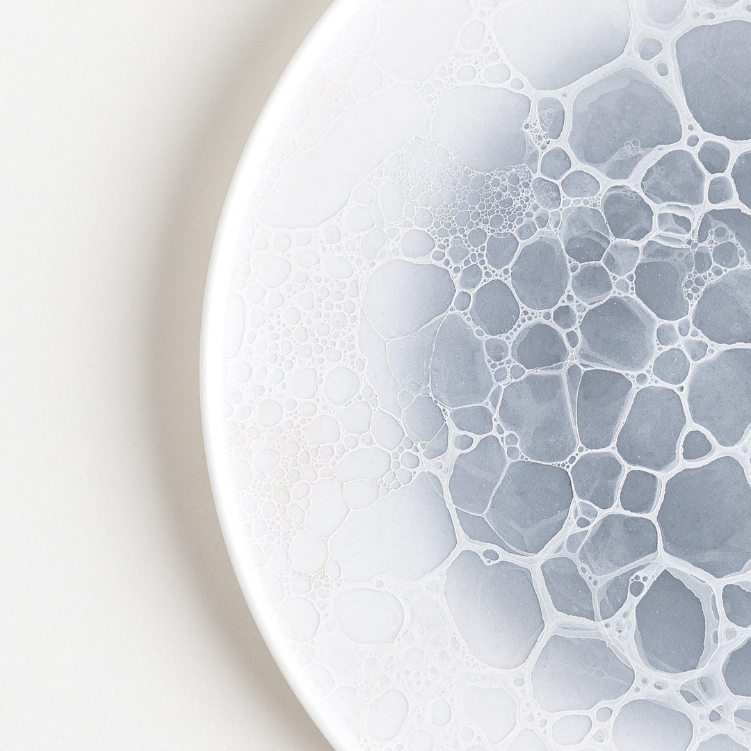 A white dinner plate featuring a unique bubble design in shades of blue, resembling frothy ocean waves.