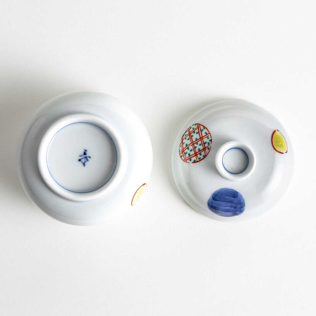 A small, shallow porcelain dish adorned with bold red and blue geometric patterns. The vibrant colors and symmetrical design create a striking contrast against the white background. This dish is perfect for serving condiments or small side dishes, reflecting the clean and precise aesthetics of Japanese tableware.