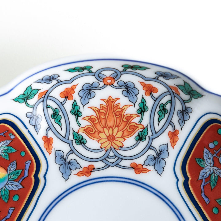 A round dessert plate featuring a central floral design with red, green, and gold patterns, bordered by sections of intricate red and green designs on the scalloped edge.