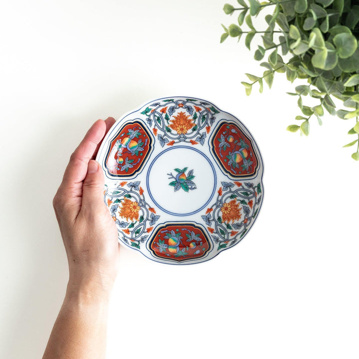 A round dessert plate featuring a central floral design with red, green, and gold patterns, bordered by sections of intricate red and green designs on the scalloped edge.