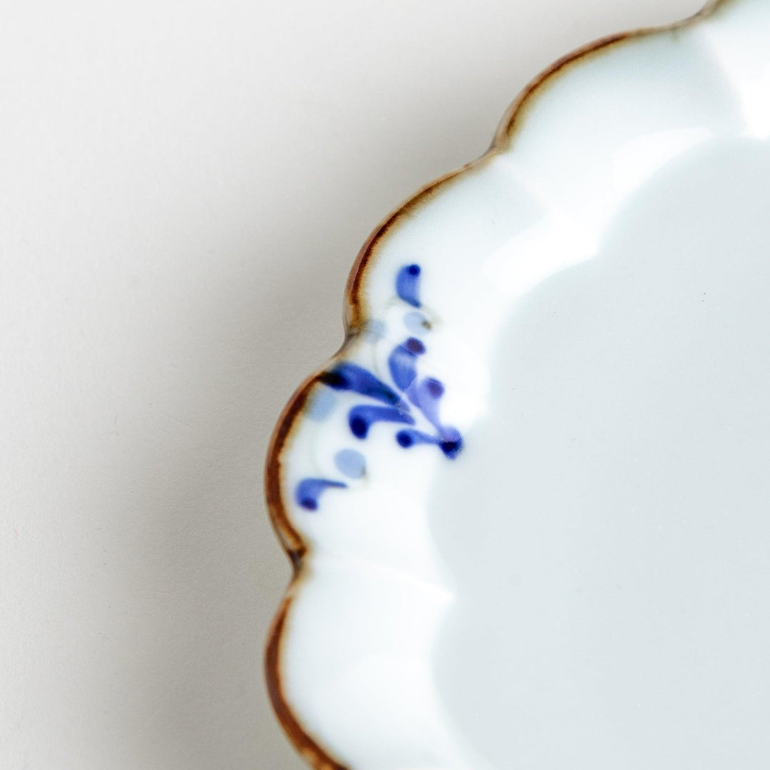 Round white porcelain dessert plate with scalloped edges, adorned with a simple blue floral pattern and brown accent trim.