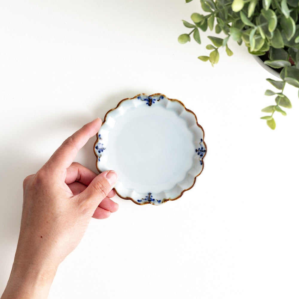 Round white porcelain sauce dish with scalloped edges, adorned with a simple blue floral pattern and brown accent trim.