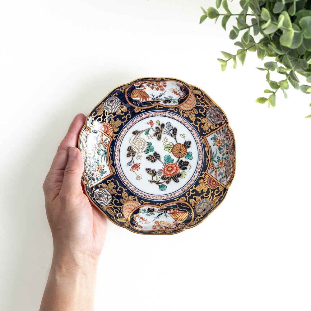 A deep round salad plate with a dark blue background, featuring a central floral design and intricate gold and red patterns with small scenes of Japanese landscapes.