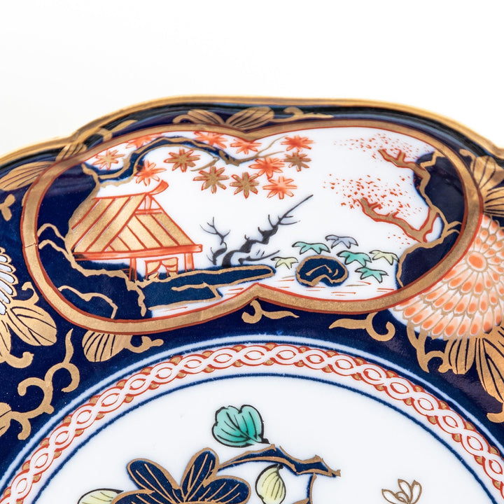 A deep round salad plate with a dark blue background, featuring a central floral design and intricate gold and red patterns with small scenes of Japanese landscapes.