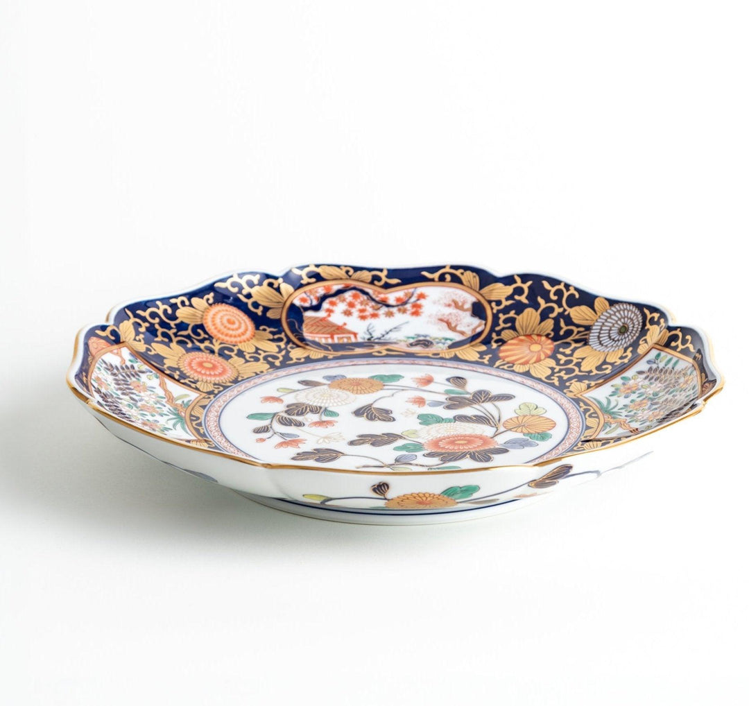 A large round serving plate with a dark blue background, featuring a central floral design surrounded by intricate gold, red, and green patterns and small scenes of Japanese landscapes.