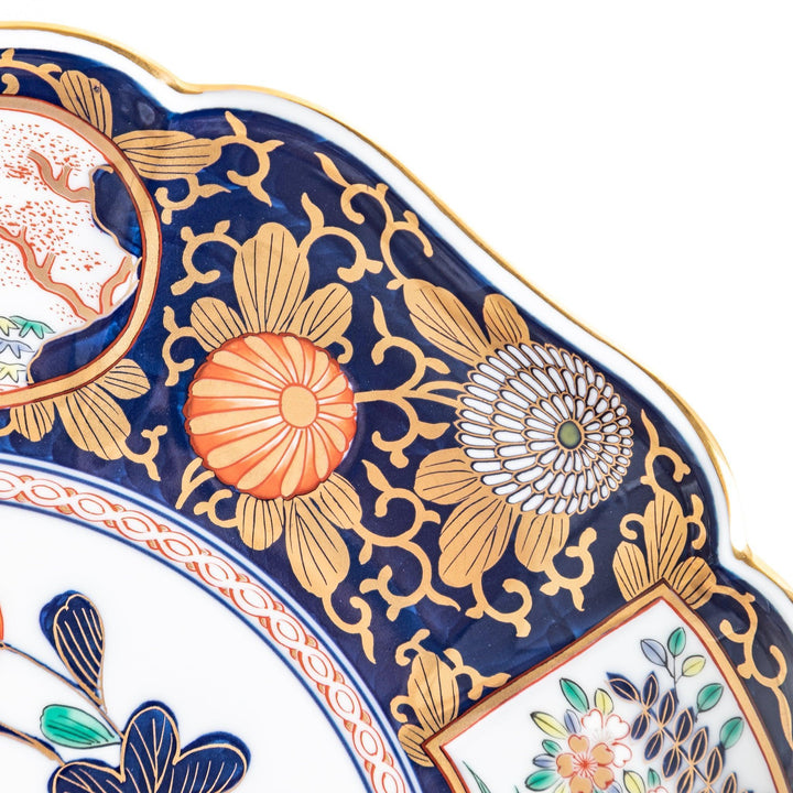 A large round serving plate with a dark blue background, featuring a central floral design surrounded by intricate gold, red, and green patterns and small scenes of Japanese landscapes.