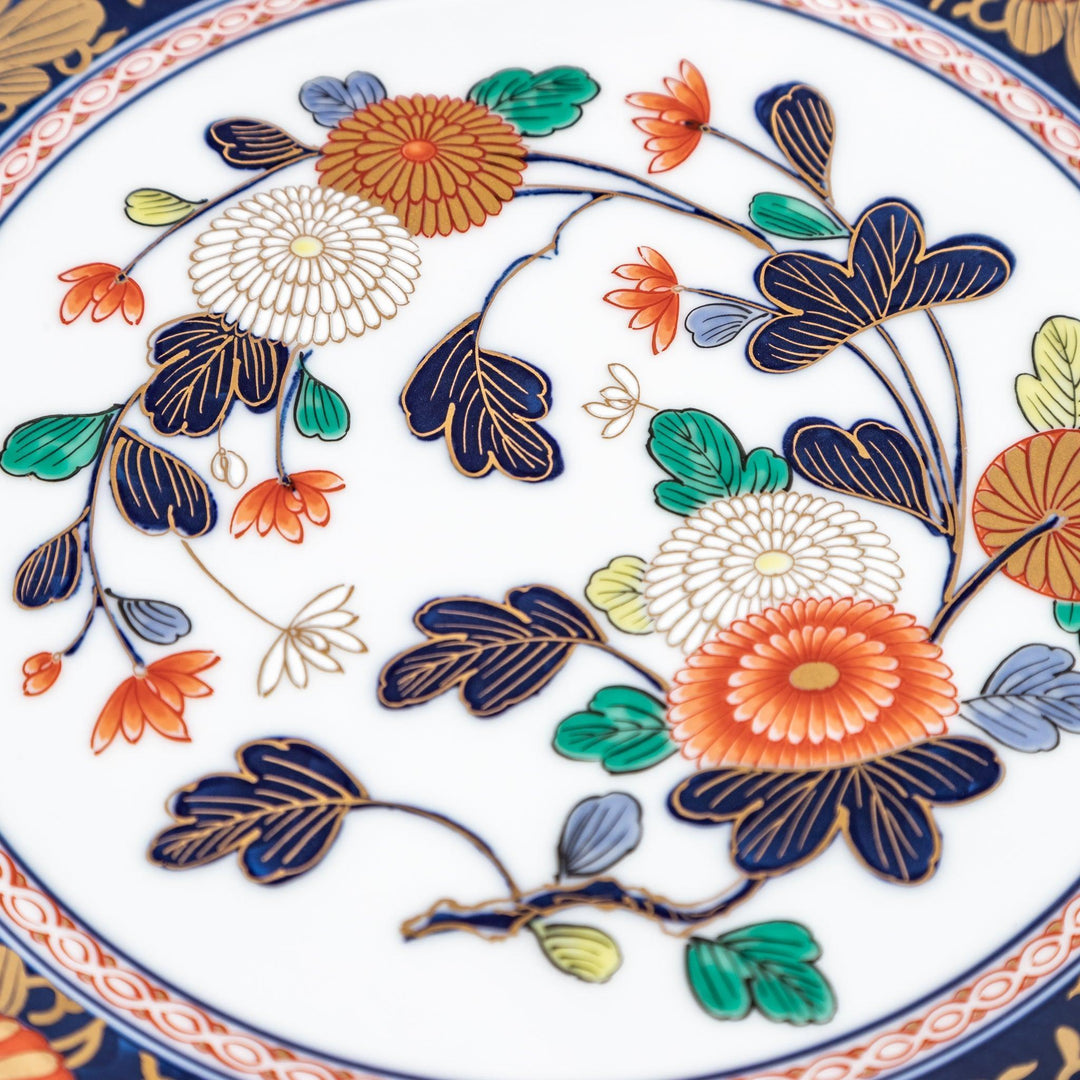 A large round serving plate with a dark blue background, featuring a central floral design surrounded by intricate gold, red, and green patterns and small scenes of Japanese landscapes.