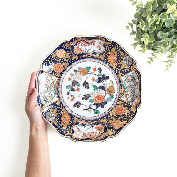 A large round serving plate with a dark blue background, featuring a central floral design surrounded by intricate gold, red, and green patterns and small scenes of Japanese landscapes.