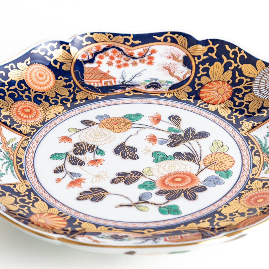 A large round serving plate with a dark blue background, featuring a central floral design surrounded by intricate gold, red, and green patterns and small scenes of Japanese landscapes.