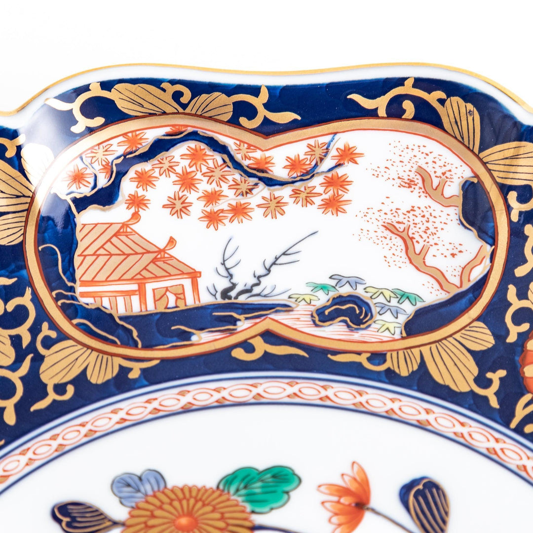 A large round serving plate with a dark blue background, featuring a central floral design surrounded by intricate gold, red, and green patterns and small scenes of Japanese landscapes.