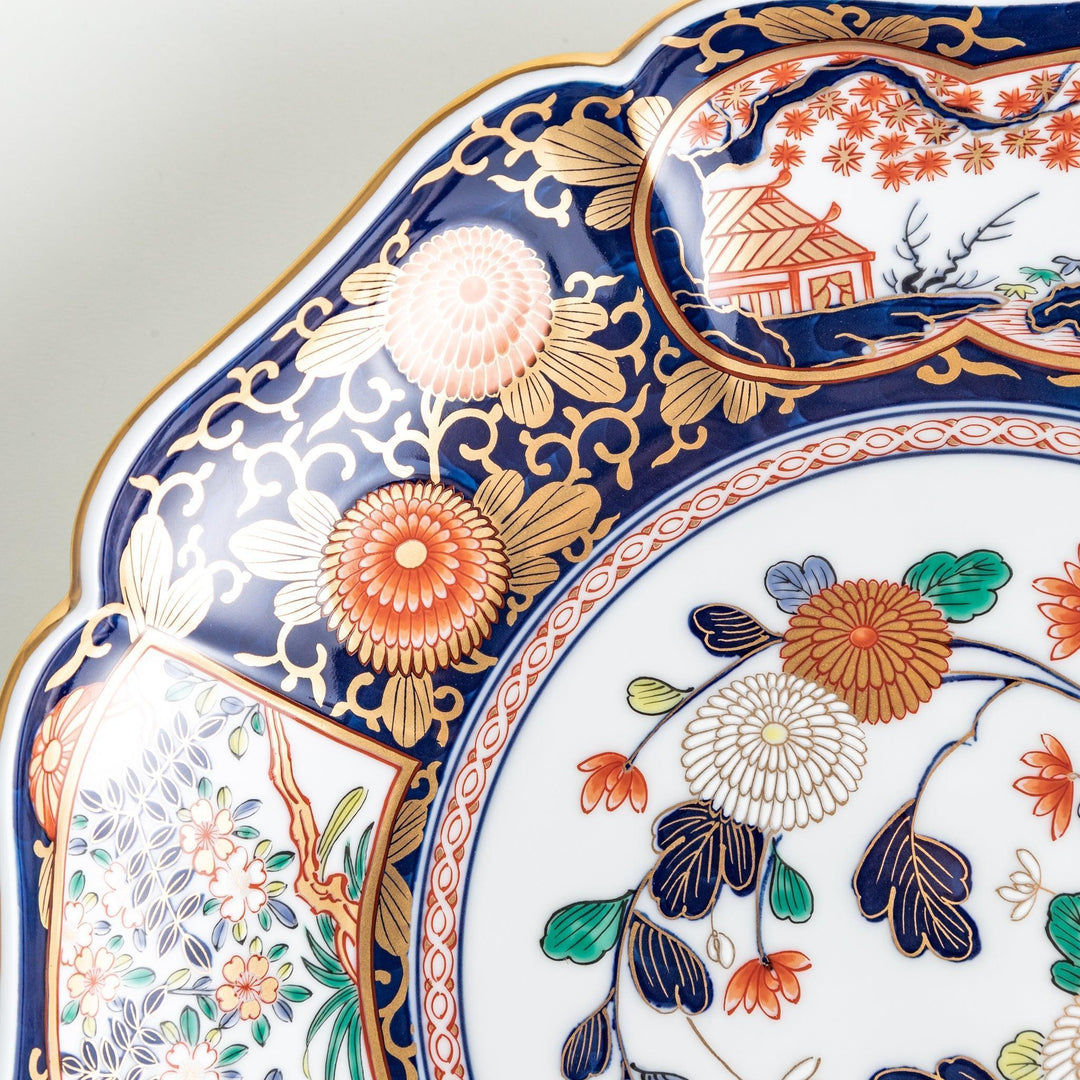 A large round serving plate with a dark blue background, featuring a central floral design surrounded by intricate gold, red, and green patterns and small scenes of Japanese landscapes.