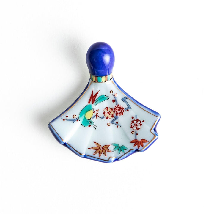 A fan-shaped ceramic chopstick rest decorated with a bird, flowers, and leaves in vibrant colors, with a blue handle.