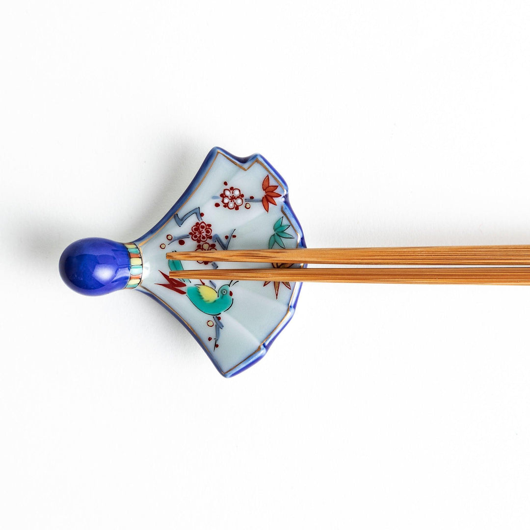 A fan-shaped ceramic chopstick rest decorated with a bird, flowers, and leaves in vibrant colors, with a blue handle.