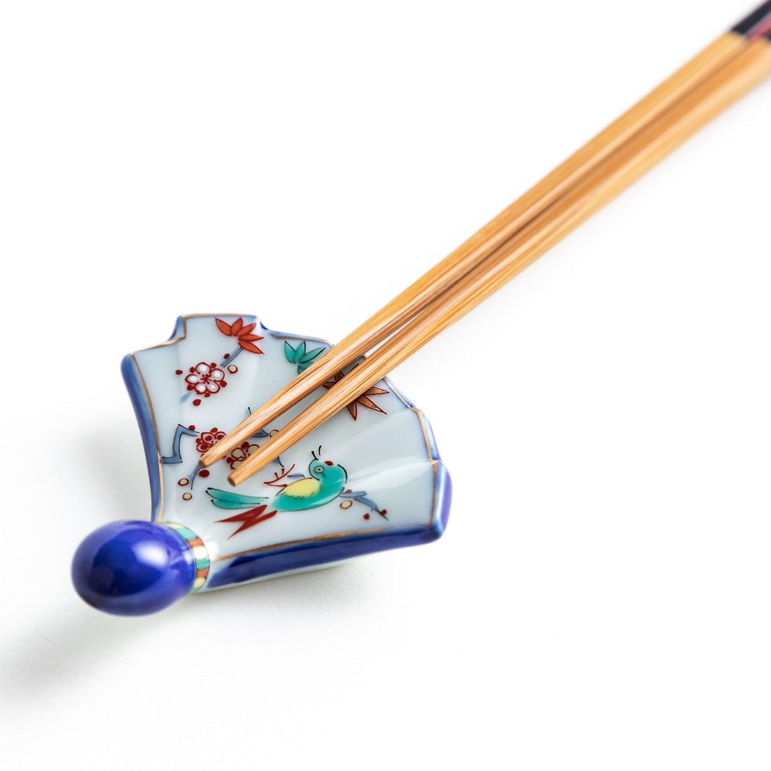 A fan-shaped ceramic chopstick rest decorated with a bird, flowers, and leaves in vibrant colors, with a blue handle.