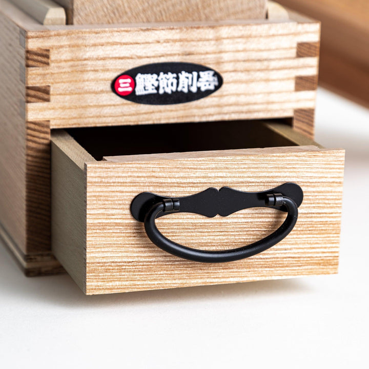 A large wooden box with a sliding drawer and lid, designed for shaving katsuobushi (dried bonito), with a colorful packaging box featuring traditional Japanese artwork.