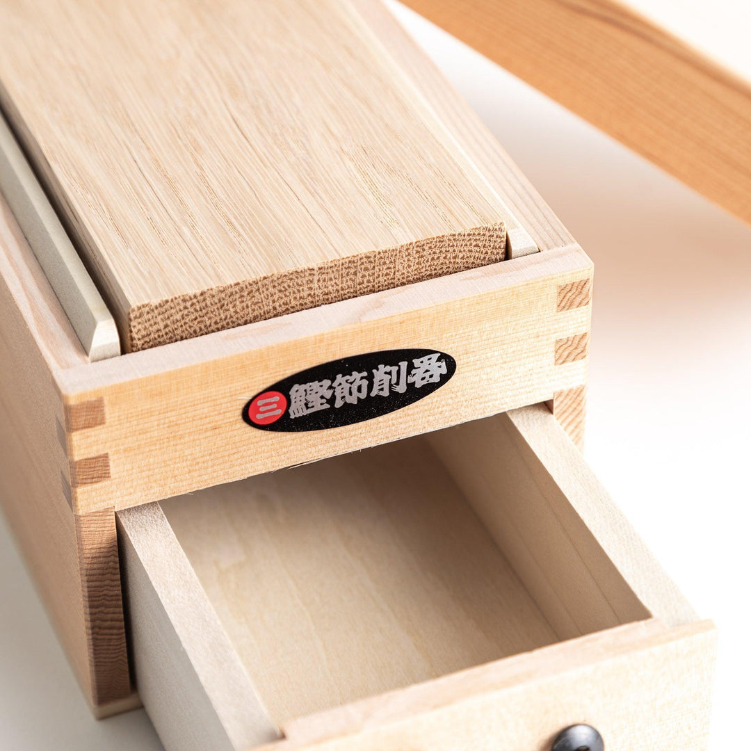 A medium-sized wooden box with a sliding drawer and lid, designed for shaving katsuobushi (dried bonito), with a colorful packaging box featuring traditional Japanese artwork.