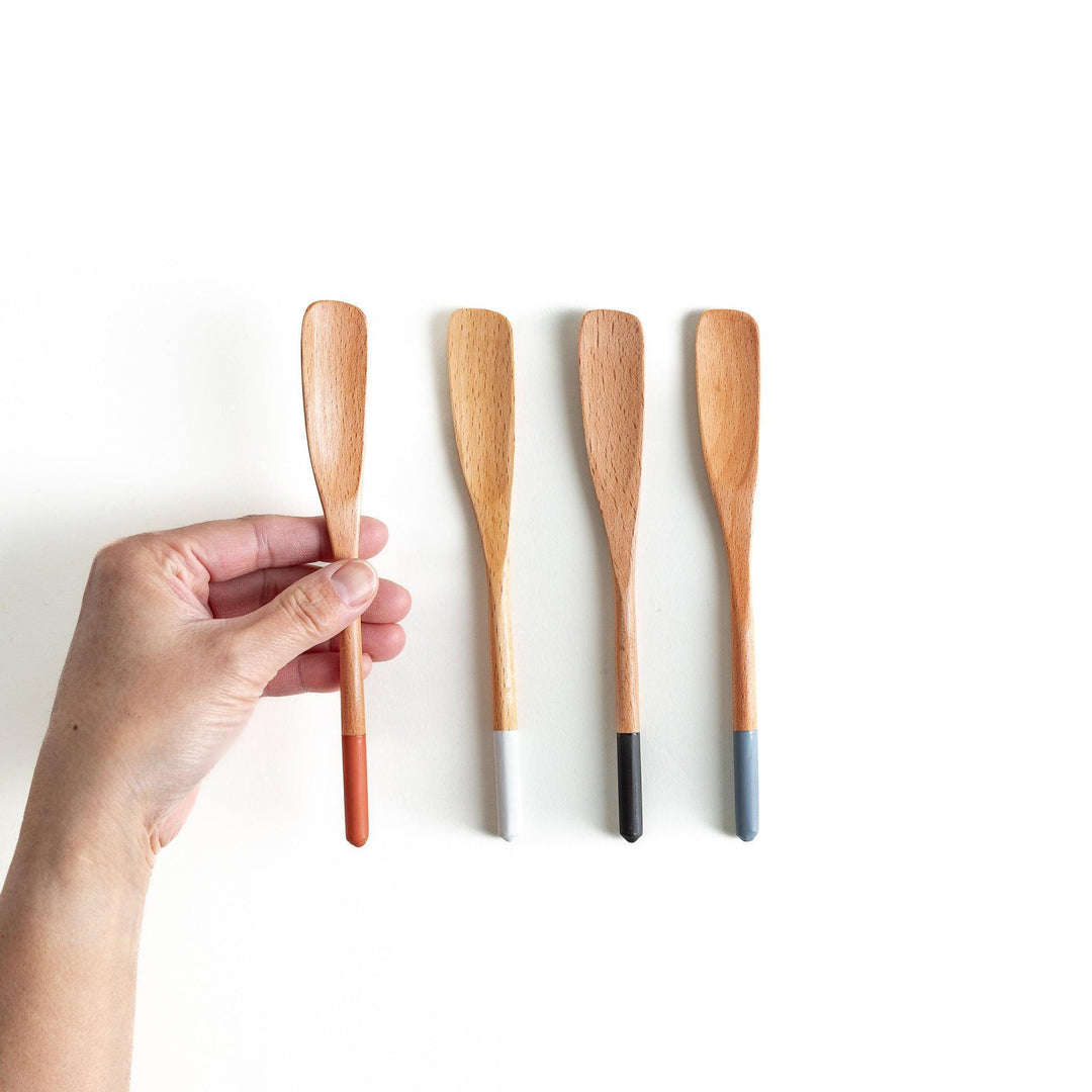 A set of four wooden spatulas with flat tips, each featuring a different colored end—white, black, blue, and red