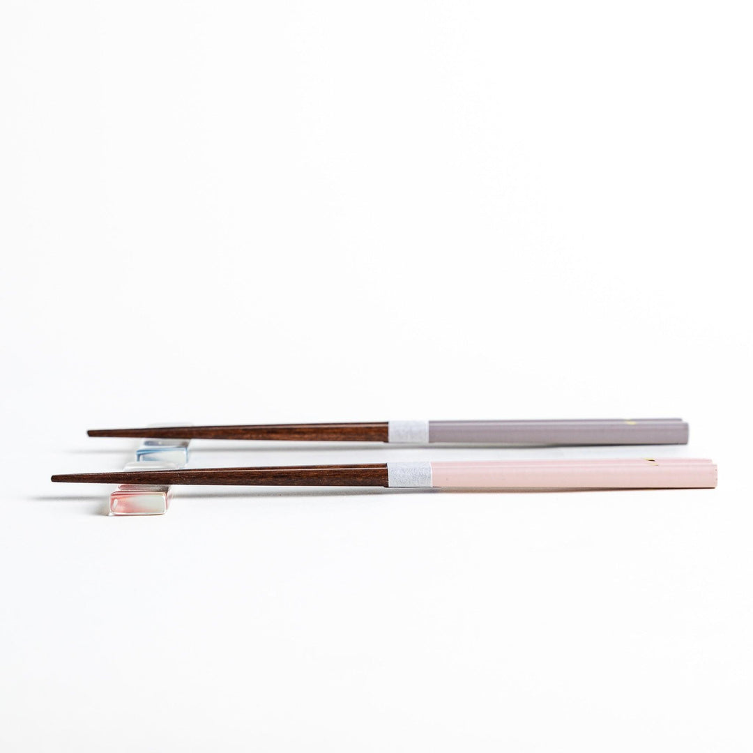 Set of two pairs of chopsticks with wooden handles, one pair in a pale gray color and the other in a light pink, presented with glass chopstick rests in a wooden gift box.