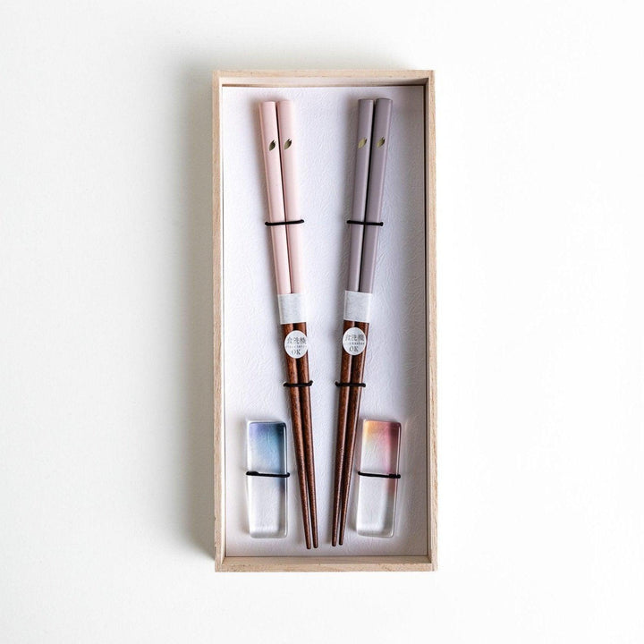 Set of two pairs of chopsticks with wooden handles, one pair in a pale gray color and the other in a light pink, presented with glass chopstick rests in a wooden gift box.