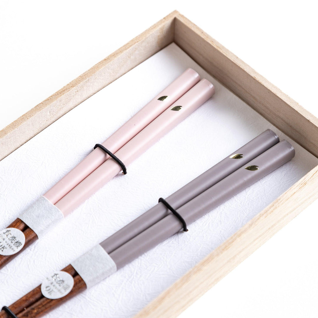 Set of two pairs of chopsticks with wooden handles, one pair in a pale gray color and the other in a light pink, presented with glass chopstick rests in a wooden gift box.