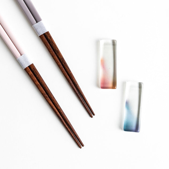 Set of two pairs of chopsticks with wooden handles, one pair in a pale gray color and the other in a light pink, presented with glass chopstick rests in a wooden gift box.