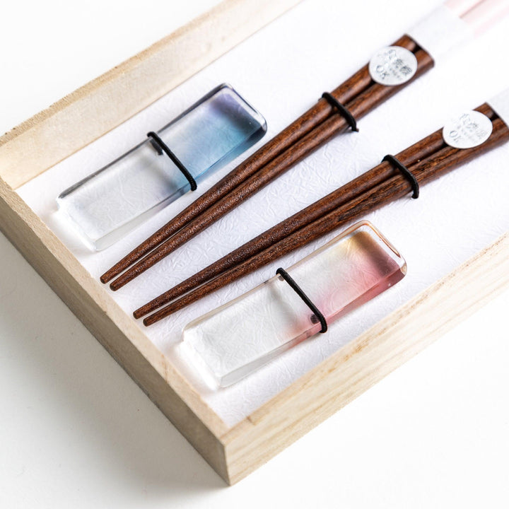 Set of two pairs of chopsticks with wooden handles, one pair in a pale gray color and the other in a light pink, presented with glass chopstick rests in a wooden gift box.