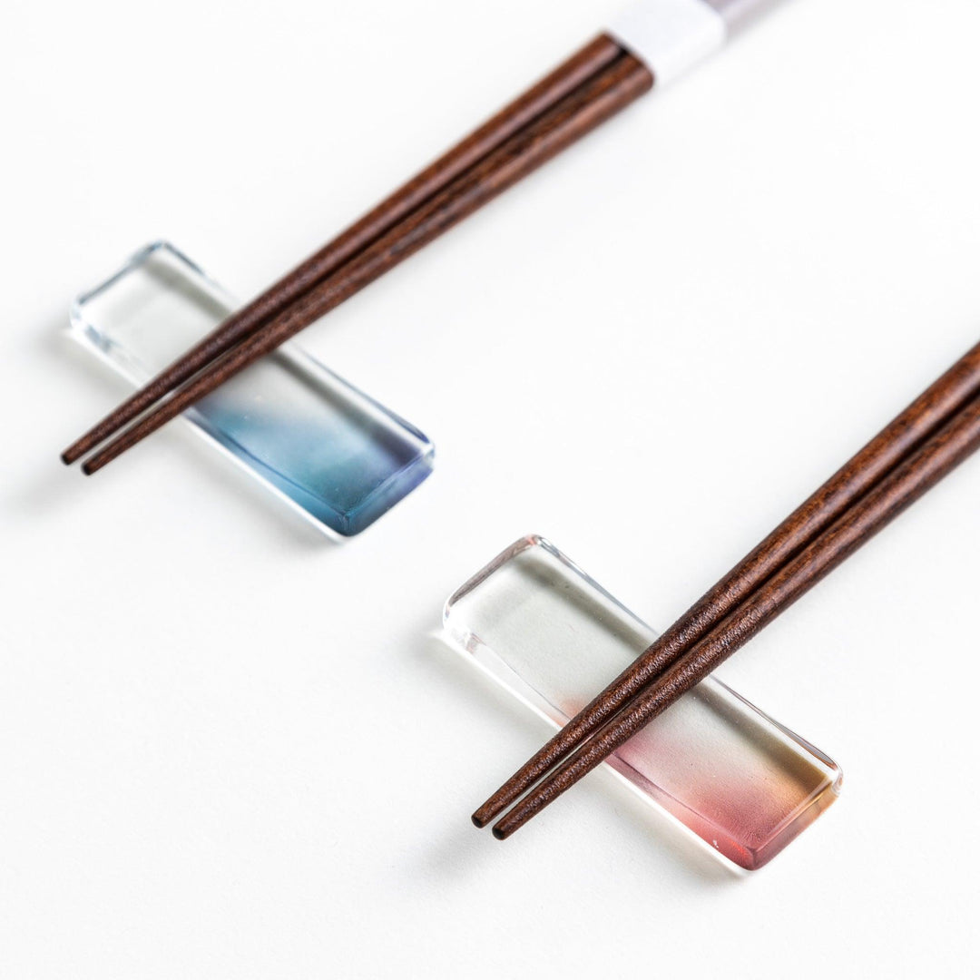 Set of two pairs of chopsticks with wooden handles, one pair in a pale gray color and the other in a light pink, presented with glass chopstick rests in a wooden gift box.