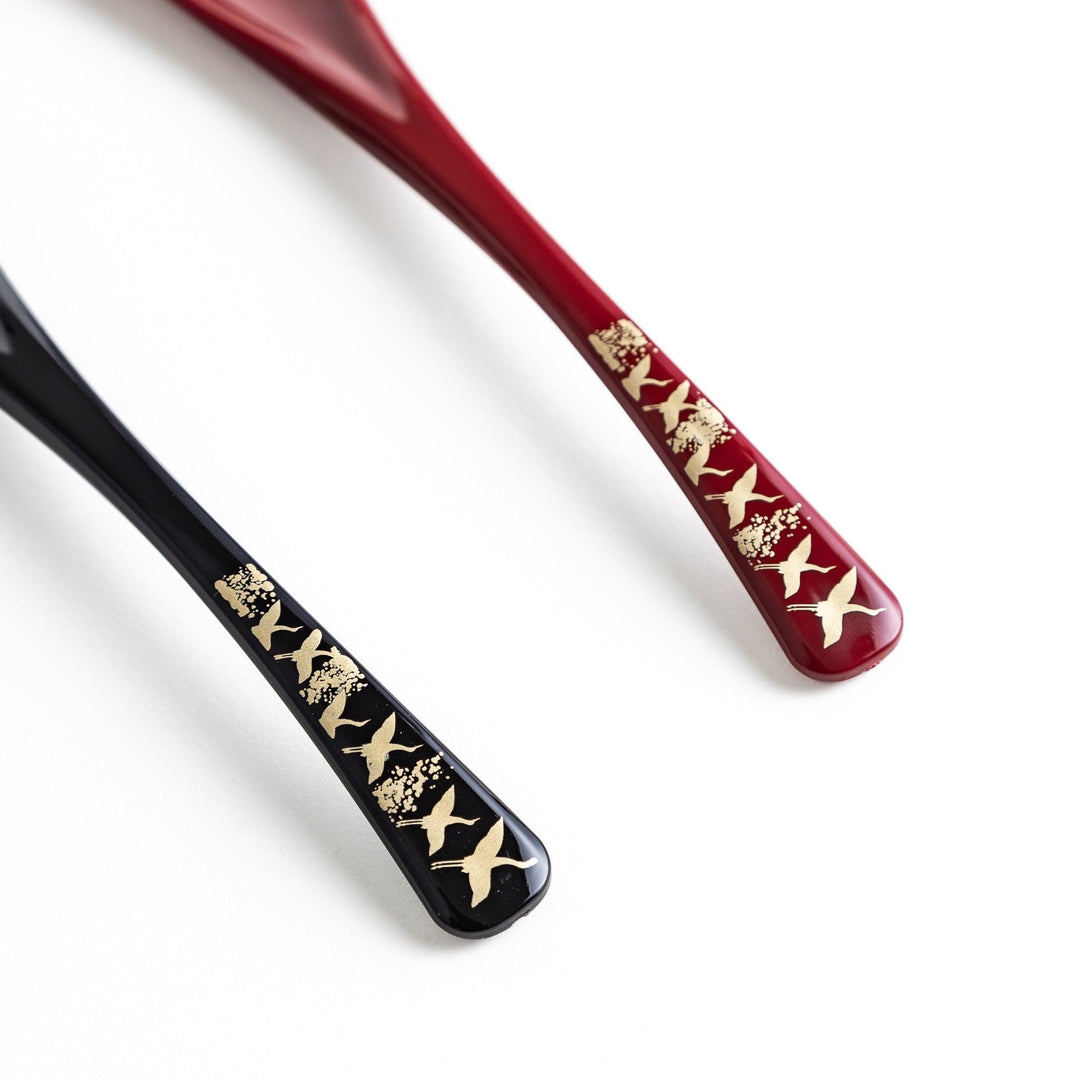 A boxed set of two lacquered spoons, one red and one black, featuring a crane pattern in gold on the handles.