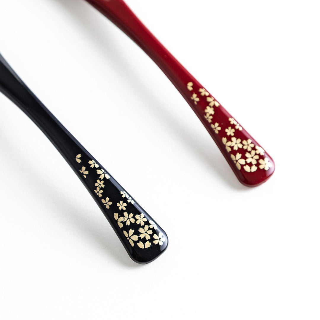 A boxed set of two lacquered spoons, one red and one black, each with gold sakura patterns on the handles.