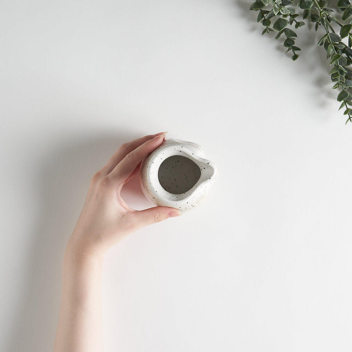 A small, off-white sauce pourer with a rounded shape, speckled glaze, and a slightly flared spout.