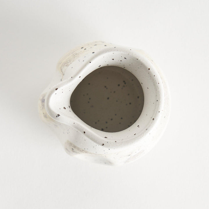 A small, off-white sauce pourer with a rounded shape, speckled glaze, and a slightly flared spout.