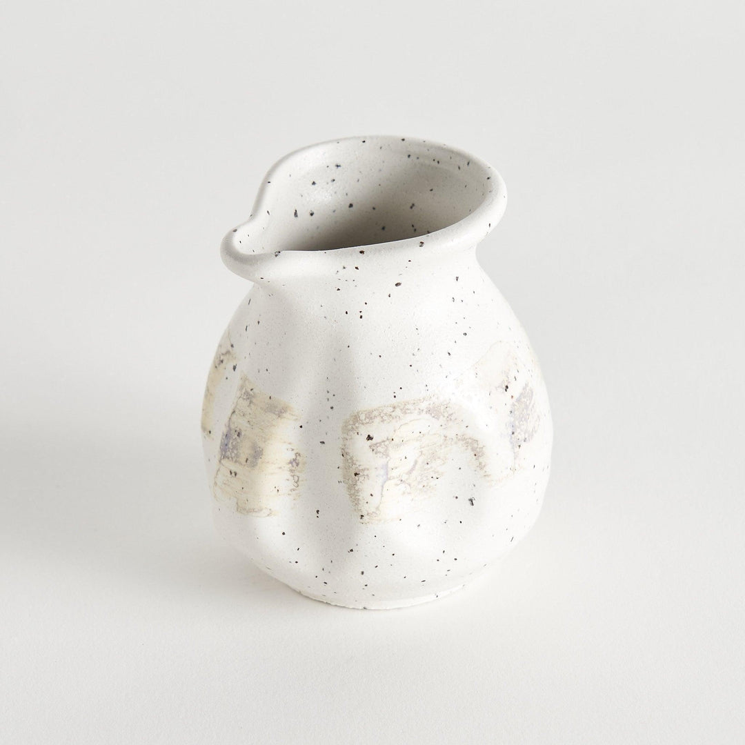 A small, off-white sauce pourer with a rounded shape, speckled glaze, and a slightly flared spout.