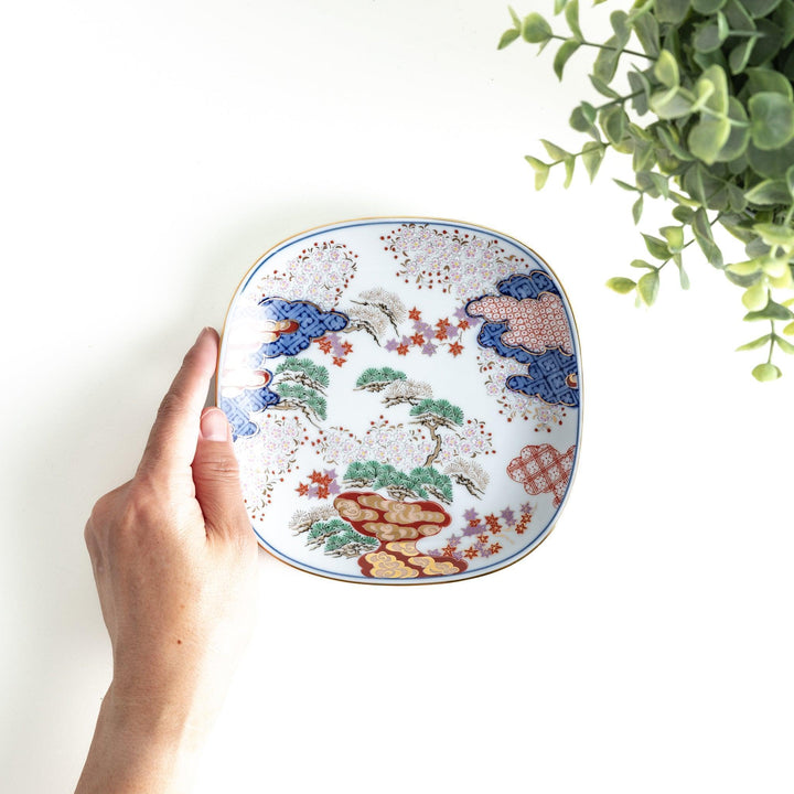 A square dessert plate featuring an intricate Japanese landscape with trees and clouds, surrounded by red and blue patterns on the border.