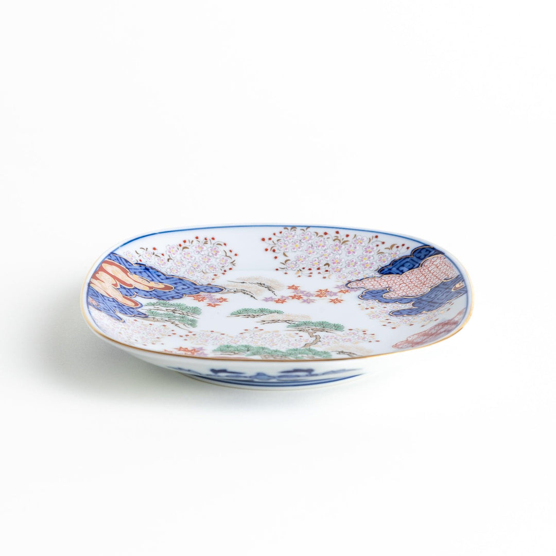 A square dessert plate featuring an intricate Japanese landscape with trees and clouds, surrounded by red and blue patterns on the border.