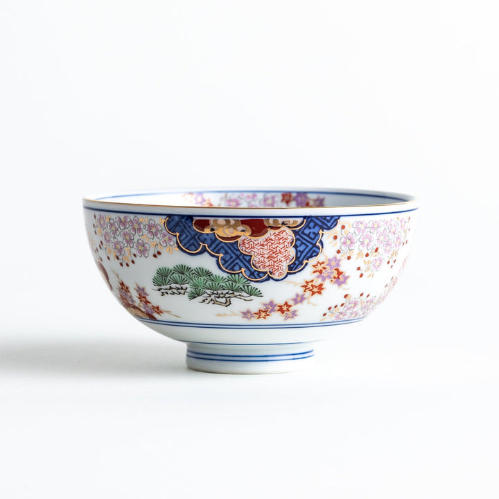 A round ceramic rice bowl with detailed traditional Japanese designs of trees and clouds in blue, red, and green on a white background.