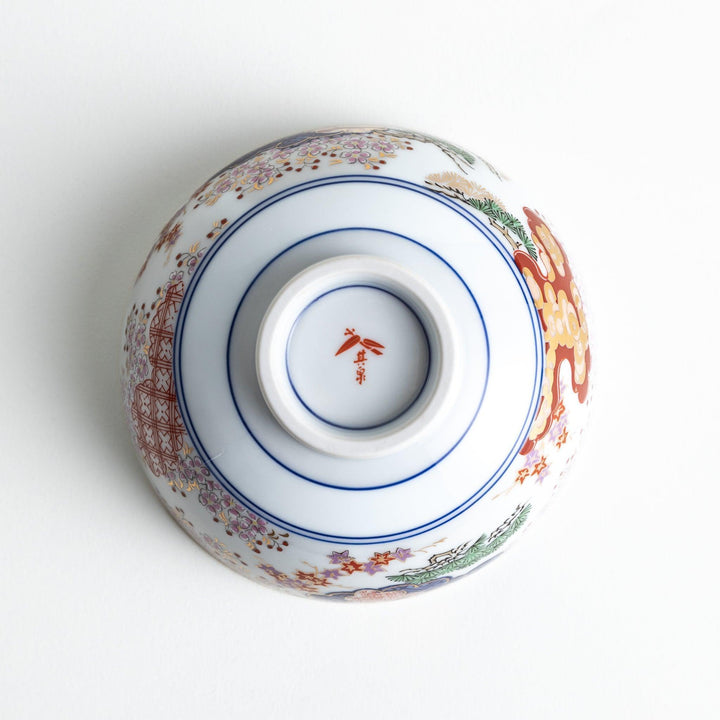 A round ceramic rice bowl with detailed traditional Japanese designs of trees and clouds in blue, red, and green on a white background.