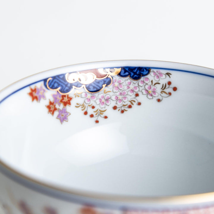 A round ceramic rice bowl with detailed traditional Japanese designs of trees and clouds in blue, red, and green on a white background.