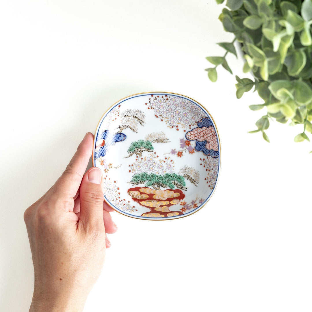 A square sauce dish featuring a traditional Japanese landscape with trees, clouds, and a golden sun, bordered by blue and red patterns.