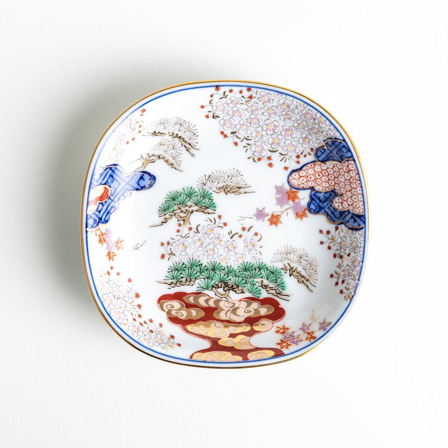 A square sauce dish featuring a traditional Japanese landscape with trees, clouds, and a golden sun, bordered by blue and red patterns.