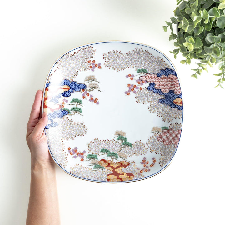 A square serving plate with a white background, decorated with a traditional Japanese landscape of trees, clouds, and red maple leaves.