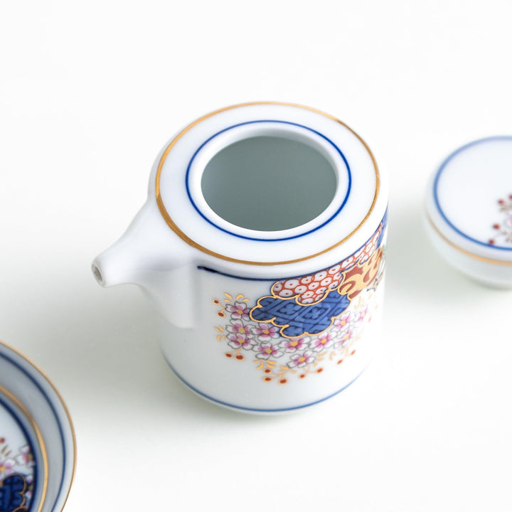 A small white sauce dispenser with intricate traditional Japanese patterns in blue, red, and gold, accompanied by a matching saucer.