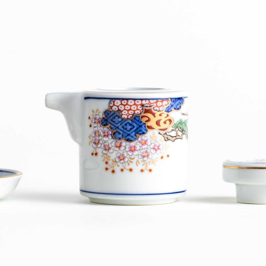 A small white sauce dispenser with intricate traditional Japanese patterns in blue, red, and gold, accompanied by a matching saucer.