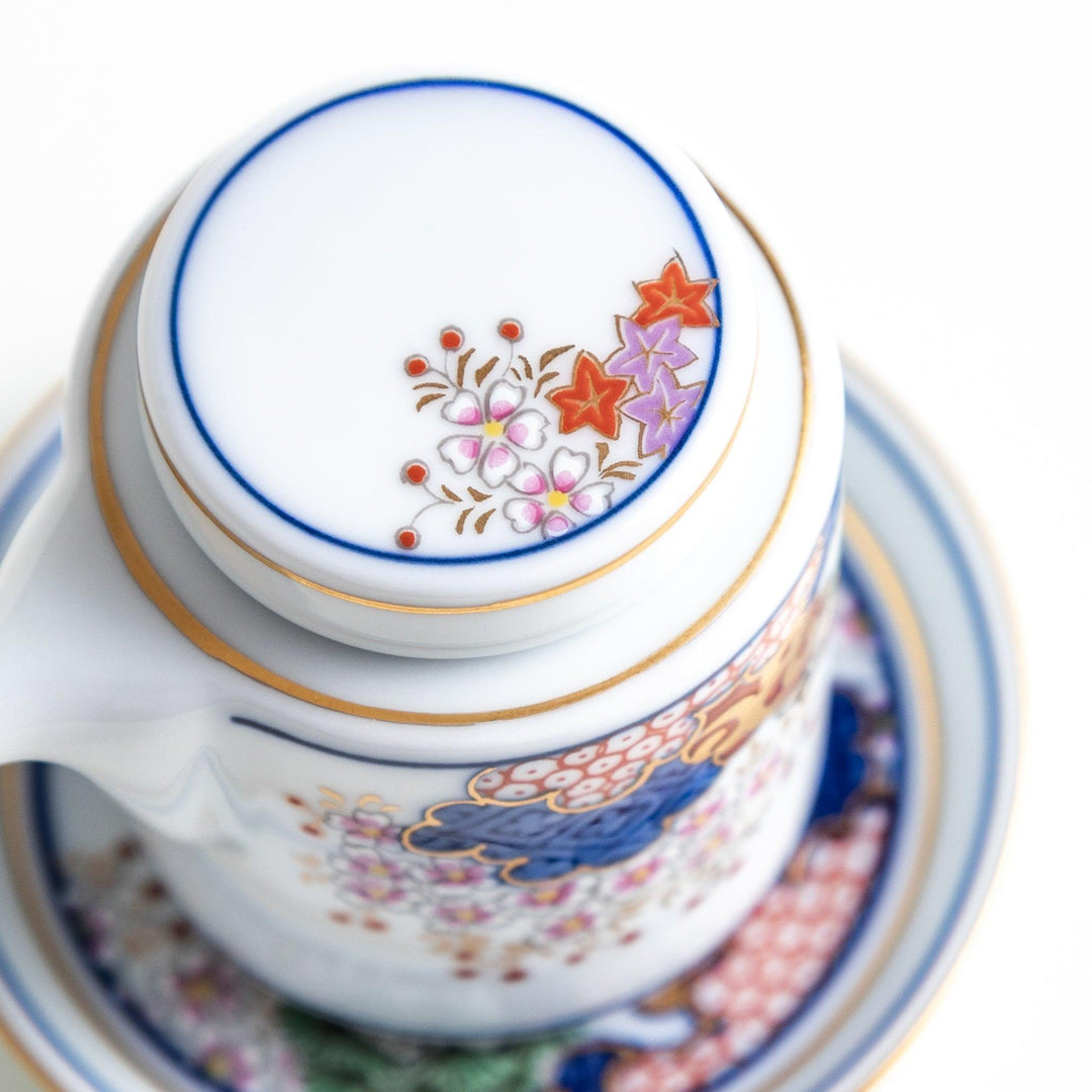 A small white sauce dispenser with intricate traditional Japanese patterns in blue, red, and gold, accompanied by a matching saucer.